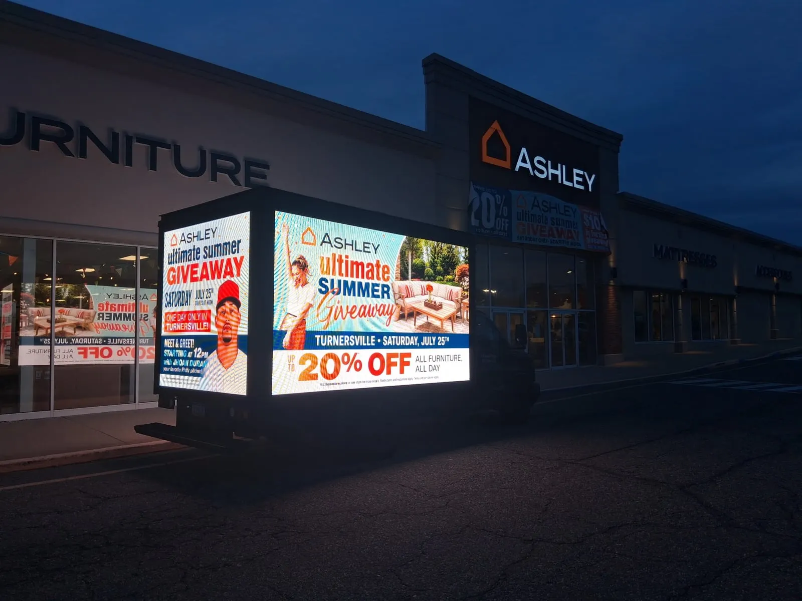 A Brandwagon LED billboard truck lit up at night outside Ashley HomeStore in Turnersville, running a summer giveaway ad