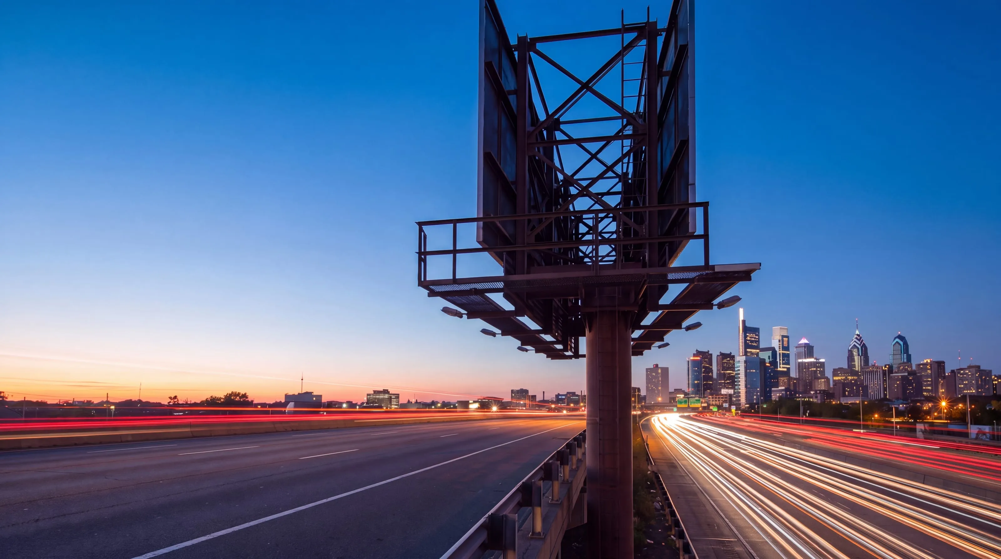 A billboard structure above I-95 traffic at dusk with the Philadelphia skyline behind it